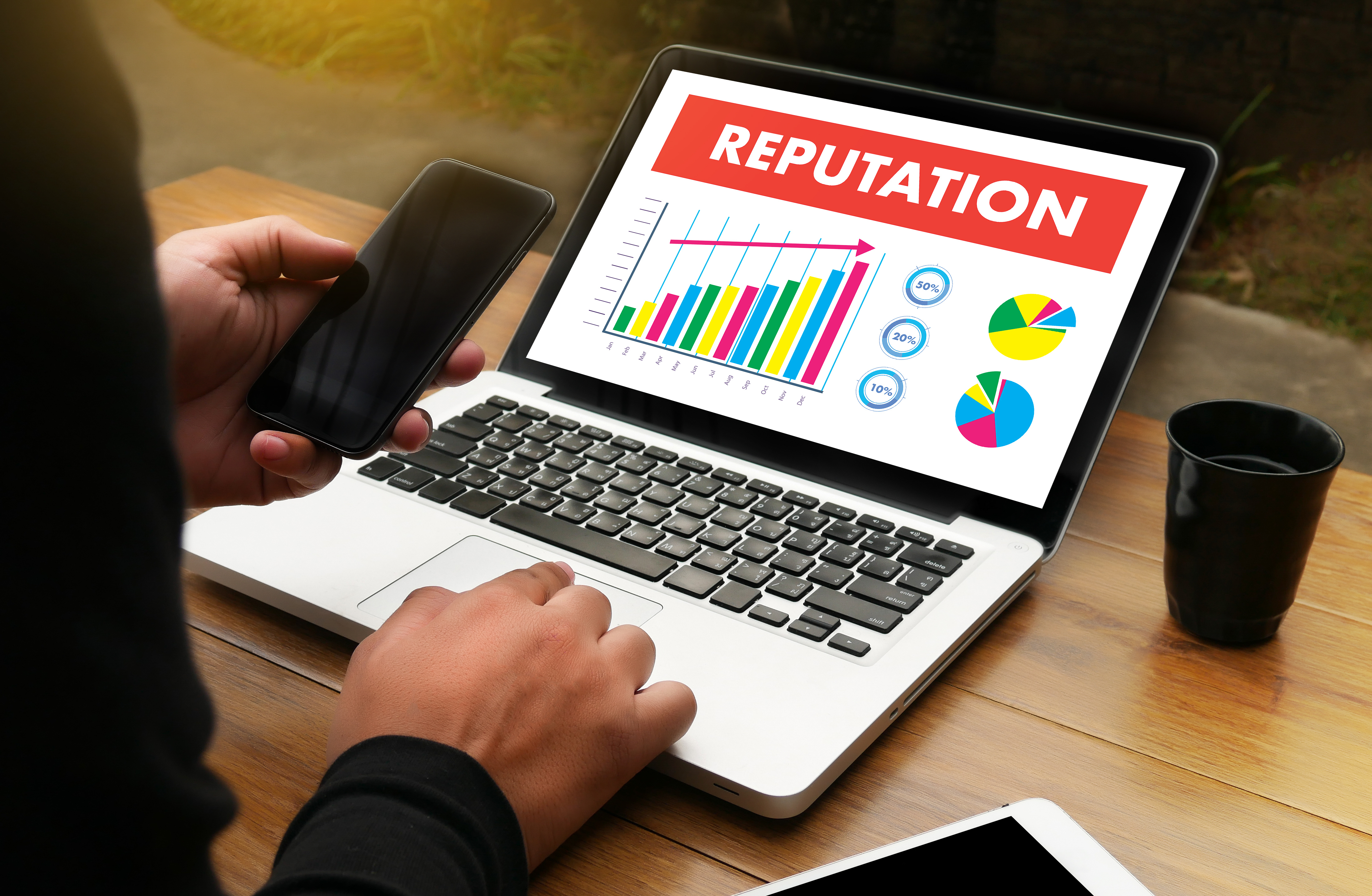 Someone sitting at a table looking at a cellphone and at a laptop showing a graph labeled reputation with a cup within reach