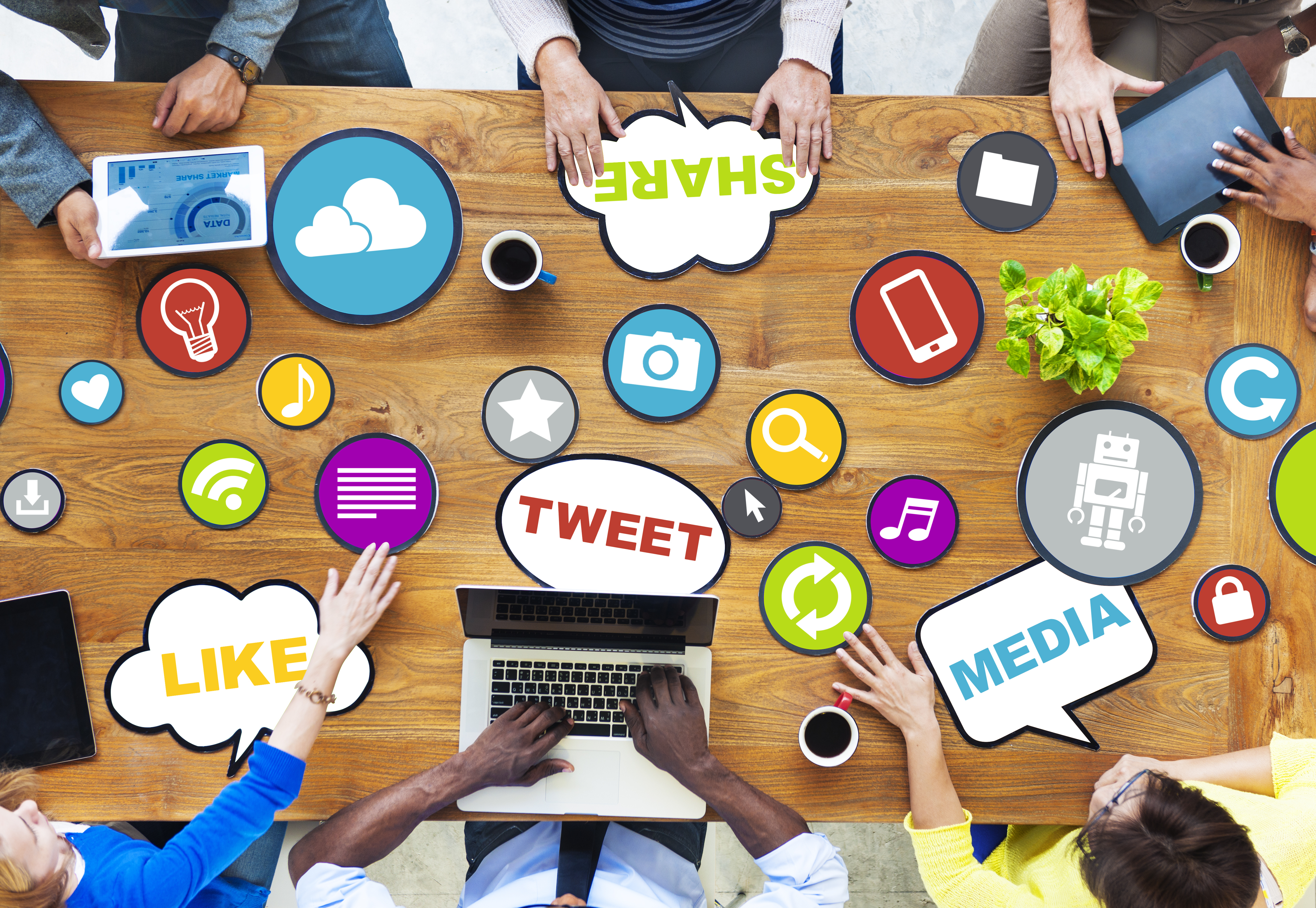 a group sitting around a table covered with symbols of social media like tweets, cloud,music each using either a laptop or a tablet with cups of coffee within reach